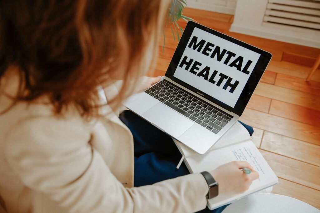A woman participating in an online therapy session, taking notes with a laptop displaying 'Mental Health'.